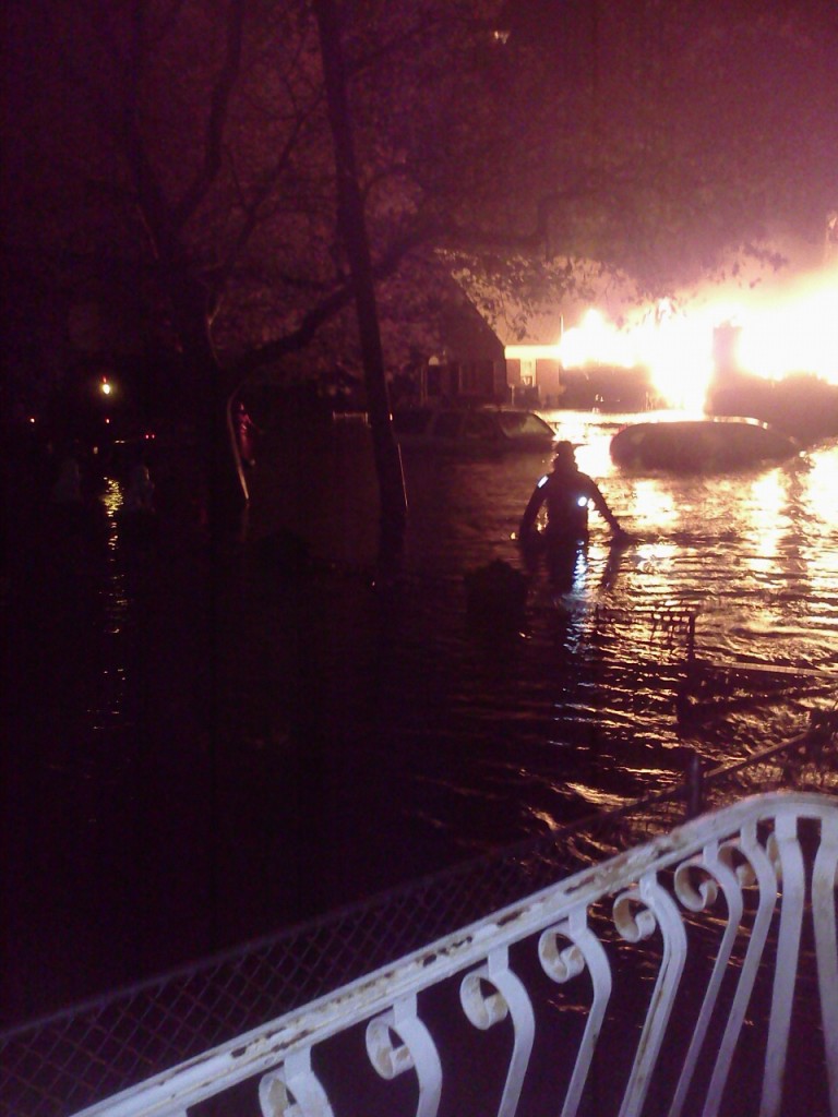 A fireman wades through the flooded street assessing the fire (next to Waldbaums in Howard Beach). The fire engine is a block away but can't get close enough to put out the fire. The car lights flicker on and off as their alarms sound and soon die off. Later, a motorboat with some elderly passengers will pass by and pick up a woman who can't swim and was stranded in her car in the middle of the flooded street. After the water recedes, the looters come. Thank God for the help of the military who were based in Waldbaums parking lot between all the flooded cars.  For the most part, things in Old Howard Beach appear to be back to normal a year after Superstorm Sandy, but it still doesn't always feel that way. –Cristie Barone Photo Courtesy Cristie Barone  
