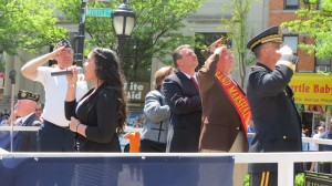 Desiree Wisotsky, center at microphone, sings the National Anthem as Master of Ceremonies Russell Goeller, left, Assemblyman Cathy Nolan, state Sen. Joe Addabbo Jr., Grand Marshal Charles Dunn, and retired U.S. Army Major General Evo Riguzzi salute the Ridgewood War Memorial, dedicated to military men and women who lost their lives in battle.  Photo by Anna Gustafson